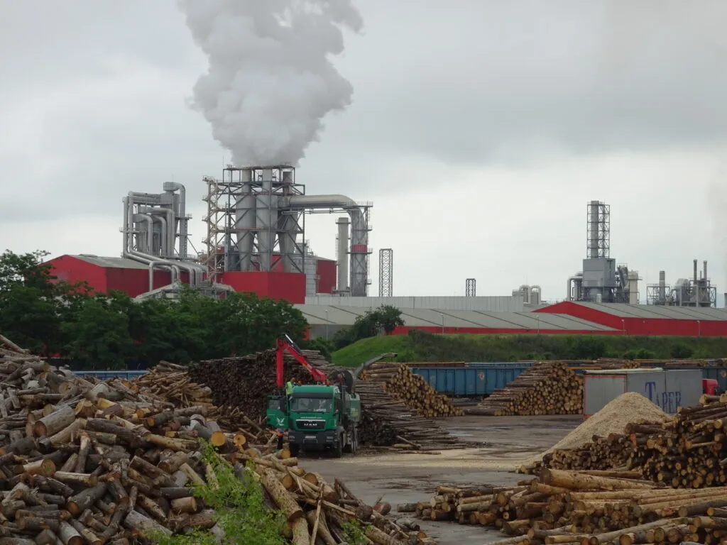 Industrieanlage mit Rauch aus einem Schornstein und Holzstapeln in der Nähe.