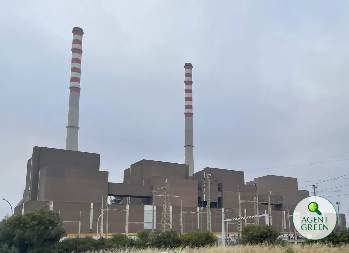 Industrieanlage mit zwei hohen Schornsteinen unter bewölktem Himmel, mit dem Agent-Green-Logo.